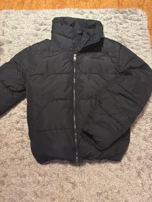 Mörkblå/svart pufferjacka  - Svart pufferjacka från Lager 157 i storlek XS. Jackan har hög krage och dragkedja framtill. Den är mycket puffig och håller värme bra. Den har ett elastiskt band runt midjan vilket ger en snatched look. Det är samma band i slutet av ärmarna. Jackan har stora fickor med knappar. Den har ett litet hål inuti jackan vid lappen vilket inte syns.