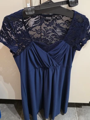 Marinblå spetsklänning Bodyflirt - Säljer en marinblå kortklänning från Bodyflirt med v-ringad omlottfront och vackra spetsdetaljer upptill och på ärmarna. Klänningen har korta ärmar och ett mjukt, fallande tyg som ger en snygg siluett. Perfekt för dig som gillar romantiska detaljer och stilren look.
