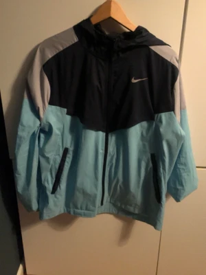 Nike vindjacka i blått och svart - Nike vindjacka med huva i färgblock av ljusblått, svart och grått. Jackan har dragkedja framtill, två fickor med dragkedja och meshfoder. Perfekt för löpning eller utomhusaktiviteter. Snygg och sportig design med Nike-logga på bröstet.