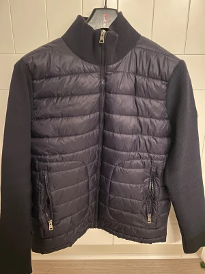 Moncler cardigan  - Säljer en mörkblå pufferjacka från Moncler med stickade ärmar och krage. Jackan har quiltad front, två fickor med dragkedja och klassisk Moncler-logga på ärmen. Insidan har ett coolt serietryck. Perfekt för dig som vill ha något både snyggt och funktionellt.