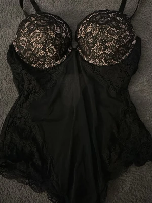 Svart spetsbody från H&M - Säljer en svart body från H&M i storlek 42 med vadderade kupor och snygga spetsdetaljer över bysten och längs sidorna. Bodyn har justerbara axelband och en elegant, feminin look med genombruten spets upptill. 