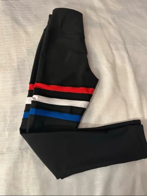 Lismina Svarta leggings Turkey med färgglada ränder - Snygga svarta leggings med bred midja och färgglada ränder i rött, vitt, blått och svart runt låren. Stretchigt material som sitter tight och är perfekt för träning eller chill. Ränderna ger en sportig vibe och gör dem till ett statement-plagg.