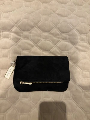 Svart mocka kuvertväska med kedja - Snygg svart kuvertväska i mocka från ” Festive Musthaves by Kim Kötter” . Väskan har en guldfärgad kedja som axelband och en dragkedja framtill. Perfekt storlek för det viktigaste och stilren design som passar till många looks.
