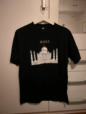 Svart t-shirt med Taj Mahal tryck - Svart t-shirt i bomull med ett stort tryck av Taj Mahal och texten 'INDIA' i guld på bröstet. Klassisk rund hals och korta ärmar. Perfekt för dig som gillar unika motiv och vill sticka ut lite extra.