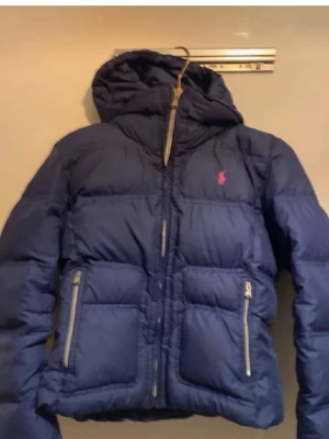 Blå pufferjacka från Polo Ralph Lauren XL - Blå pufferjacka från Polo Ralph Lauren i storlek XL i barn men passar S dam . Jackan har huva, två stora fickor med dragkedja och guldfärgade detaljer. Klassisk röd Polo-logga på bröstet och quiltad design. Perfekt för kalla dagar och riktigt snygg stil.