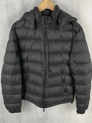 Svart pufferjacka från Massimo Dutti - Snygg svart pufferjacka från Massimo Dutti med huva och dragkedja framtill. Jackan har två sidofickor med dragkedja och justerbara muddar med knapp. Perfekt för kalla dagar och har en modern, stilren look.