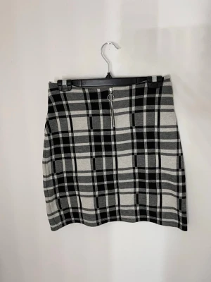 Rutig kortkjol med dragkedja - Säljer en snygg rutig kortkjol i svart, grått och vitt med en dekorativ dragkedja framtill. Den är egentligen 164 barnstorlek men passar mig som har storlek 38. Kjolen har en tight elastisk passform. 