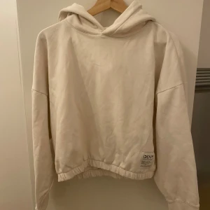Beige hoodie från Denim - Säljer en beige hoodie från Denim med avslappnad passform och elastisk nederkant. Hoodien har en klassisk huva och en liten etikett med logga nedtill. Tillverkad i mjukt bomullsmaterial, perfekt för en chill och enkel stil.