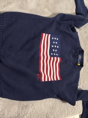 Mörkblå stickad tröja Polo Ralph Lauren - Säljer en mörkblå stickad tröja från Polo Ralph Lauren med amerikansk flagga i rött, vitt och blått på bröstet. Tröjan har rund halsringning och ribbade muddar. RL-broderi i rött nedanför flaggan. Perfekt för dig som gillar klassisk college-stil.