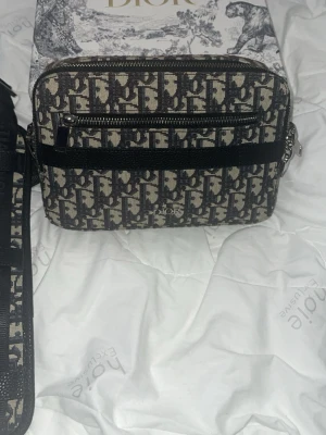 Dior axelväska med monogrammönster - Säljer en snygg axelväska från Dior med klassiskt monogrammönster i svart och beige. Väskan har justerbar axelrem, dragkedja och en praktisk ytterficka. Perfekt för dig som vill ha en trendig och lyxig accessoar till din outfit.