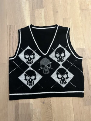 Svart väst med dödskallemönster - Stickad svart väst med vita och grå dödskallar i argyle-mönster. Västen har V-ringning och vita detaljer vid hals och nederkant. Perfekt för dig som gillar alternativ stil och vill sticka ut.