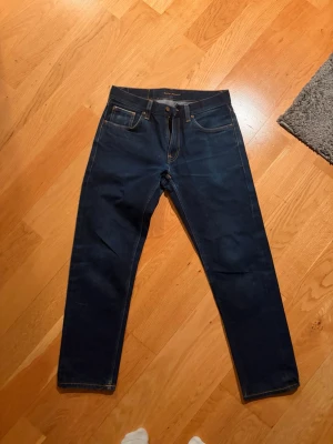 Mörkblå jeans från Nudie Jeans - Säljer ett par mörkblå jeans från Nudie Jeans med klassisk femficksmodell och raka ben. Jeansen har kontrastsömmar och Nudie-logga på bakfickan. Perfekta för dig som gillar stilrena och tidlösa jeans. Modellen Gritty Jackson 