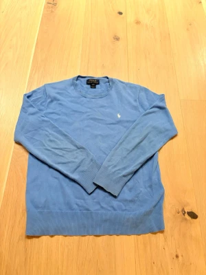 Blå stickad tröja från Polo Ralph Lauren - Klassisk blå stickad tröja från Polo Ralph Lauren med rund halsringning och diskret broderad logga på bröstet. Tröjan har långa ärmar och ribbade muddar vid ärmslut och nederkant. Perfekt för lager på lager och enkel att matcha. Tveka inte på att höra av dig vid minsta fråga😊