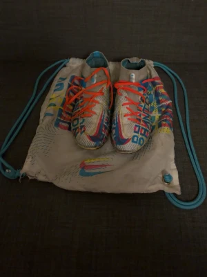 Färgglada Adidas fotbollsskor - Säljer ett par riktigt färgglada Adidas fotbollsskor med mönster i blått, rött och gult på vit bas. Skorna har snörning med orangea skosnören och är tillverkade i syntetmaterial. Perfekta för dig som vill sticka ut på planen! Skriv vid fundering 