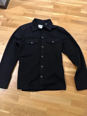 Svart overshirt från Jack & Jones - Säljer en svart overshirt från Jack & Jones med klassisk krage och knäppning framtill. Två bröstfickor med lock och knappar ger en clean look. Perfekt att bära över en t-shirt eller hoodie för en avslappnad stil.