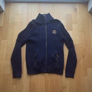 Marinblå kabelstickad tröja Lauren - Snygg marinblå kabelstickad tröja från Lauren Ralph Lauren med dragkedja framtill, två fickor med guldfärgade dragkedjor och broderad logga på bröstet. Hög ribbad krage och klassisk preppy stil. Perfekt för dig som gillar tidlös design.