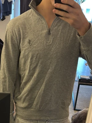 Grå halvzip från Polo Ralph Lauren - Snygg grå långärmad tröja från Polo Ralph Lauren med halvzip och hög krage. Tröjan har det klassiska broderade logotypen på bröstet och är gjord i mjukt bomullsmaterial. Perfekt för en clean och avslappnad stil.