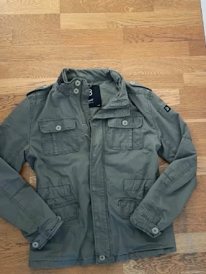 Olivgrön field jacket från Brandit  - Snygg grön utilityjacka från Brandit med fyra framfickor, axelklaffar och knappar. Jackan har dragkedja och knäppning framtill samt detaljerade sömmar. Perfekt för dig som gillar en avslappnad och cool stil. OBS - Saknar en knapp