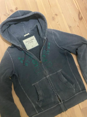 Blå hoodie från Abercrombie & Fitch - Mysig blå hoodie från Abercrombie & Fitch med dragkedja, huva med dragsko och grönt tryck på bröstet. Två fickor framtill och mjukt material på insidan. Perfekt för en avslappnad och cool stil. Tveka inte påa tt ge bud eller fråga!