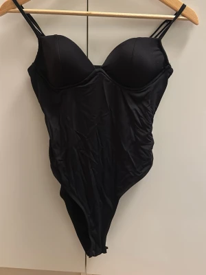 Svart baddräkt med vadderade kupor - Snygg svart baddräkt med vadderade kupor och dubbla smala axelband. Baddräkten har en enkel, stilren design och ger bra stöd upptill. Materialet är stretchigt och följsamt, perfekt för strand eller pool.