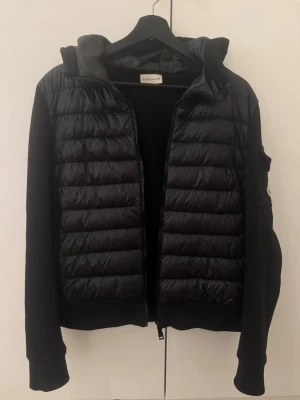 Svart dunjacka från Moncler med huva - Snygg svart dunjacka från Moncler med quiltad front, huva och dragkedja. Jackan har långa ärmar i mjukare material och en Moncler-logga på ärmen. Perfekt för kyliga dagar och ger en sportig men stilren look.