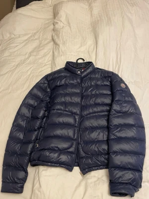 Moncler acorus jacka  - Snygg mörkblå dunjacka från Moncler med quiltad design och hög krage. Jackan har dragkedja framtill och diskret Moncler-logga på ärmen. Perfekt för kalla dagar och ger en stilren look. Stl 7 i moncler som motsvarar xl hör av er vid frågor