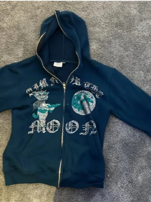 Blå hoodie Aim For The Moon XS - Mörkblå hoodie med dragkedja från Aim For The Moon. Framsidan har ett stort tryck med texten 'AIM FOR THE MOON', en tecknad katt i krona och en glittrig måne. Hoodien har huva och ribbade muddar. Perfekt för dig som gillar streetwear och coola prints.