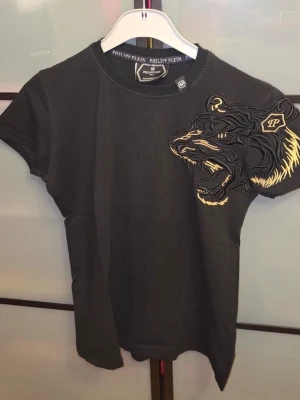 Philipp Plein T-shirt - Svart t-shirt från Philipp Plein med korta ärmar och en stor broderad tiger i svart och gult på bröstet. T-shirten har rund halsringning och är gjord i mjuk bomull. Perfekt för dig som vill sticka ut med en unik detalj. Den är använd fåtal gånger och den kommer tvättad. Tveka inte för att ställa frågor! Priset är inte hugget i sten! 