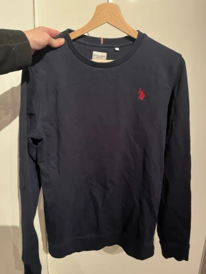Mörkblå sweatshirt från U.S. Polo Assn. - Mörkblå sweatshirt från U.S. Polo Assn. med rund halsringning och röd broderad logga på bröstet. Tröjan har långa ärmar och är gjord i mjukt bomullsmaterial. Enkel och stilren design som passar till det mesta. Storlek 14-15 år