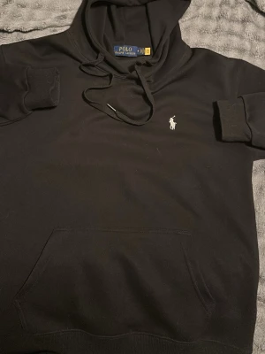 Svart hoodie från Polo Ralph Lauren - Svart hoodie från Polo Ralph Lauren med klassisk vit broderad logga på bröstet. Hoodien har huva med dragsko och en stor känguruficka framtill. Tillverkad i mjukt material som är skönt att bära. Perfekt för en avslappnad och stilren look.