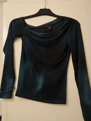 Mörkblå långärmad topp från Amisu - Snygg mörkblå långärmad topp från Amisu med draperad halsringning och figurnära passform. Tyget har en lätt skimrande effekt och toppen har en unik asymmetrisk detalj vid axeln. Ser lite ut som norrsken.