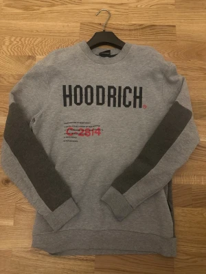 Grå sweatshirt från Hoodrich - Snygg grå sweatshirt från Hoodrich med svart tryck på bröstet och textdetaljer i svart och rött. Tröjan har mörkare grå paneler längs ärmarna och ribbade muddar. Perfekt för en streetwear-look och riktigt bekväm att bära.