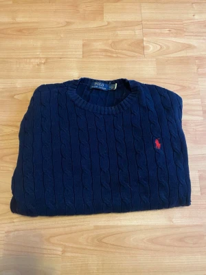 Marinblå kabelstickad tröja Polo Ralph Lauren - Kabelstickad marinblå tröja från Polo Ralph Lauren med rund halsringning och röd broderad logga på bröstet. Tröjan har långa ärmar och ribbade muddar vid ärmslut och nederkant. Perfekt för en klassisk och stilren look.