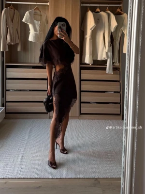 Svart asymmetrisk klänning med spets - Trendig svart klänning med asymmetrisk fåll och spetsdetaljer. Klänningen har kort ärm, markerad midja och en unik skärning med öppen detalj vid midjan. Perfekt för dig som vill sticka ut med en edgy och elegant look.