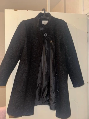 Svart kappa  - Stilren svart kappa från Isolde i storlek 36. Kappan har en rak passform, lång ärm och stängs med knappar framtill. Materialet är mjukt och har en lätt struktur som ger en snygg look. Perfekt för kyligare dagar och enkel att matcha med olika outfits.