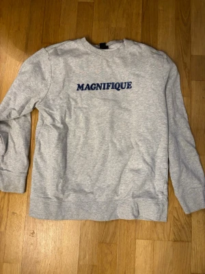 Grå sweatshirt med blå text från H&M - Säljer en ljusgrå sweatshirt från H&M i storlek S. Tröjan har trycket 'MAGNIFIQUE' i blått framtill och är långärmad med rund hals. Perfekt för en chill och avslappnad stil.