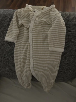 Bebis kläder Randig beige babybody  - Supersöt babybody från Cloirs i storlek 56. Den är beige och vit med smala ränder och har tryckknappar framtill för enkel påklädning. Tillverkad i mjuk bomull som känns skön mot huden. Perfekt för små bebisar!
