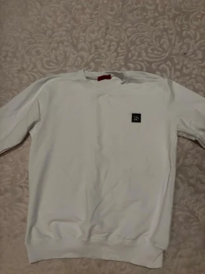 Vit sweatshirt från Hugo - Snygg vit sweatshirt från Hugo med rund halsringning och en svart patch med logga på bröstet. Tröjan har en klassisk passform och ribbade muddar vid ärmslut och nederkant. Perfekt för en clean och stilren look.