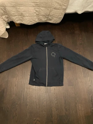 Mörkblå hoodie från Hugo Boss - Snygg mörkblå hoodie från Hugo Boss med dragkedja framtill och två fickor. Diskret tryck på bröstet och klassisk huva. Tillverkad i mjukt bomullsmaterial som känns skönt mot huden. Perfekt för en avslappnad och stilren look.