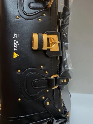 Vintage väska - Snygg svart handväska med guldfärgade detaljer och dekorativt lås framtill. Väskan har flera remmar och nitar som ger en cool look. Perfekt för dig som vill ha en trendig accessoar med plats för det viktigaste.