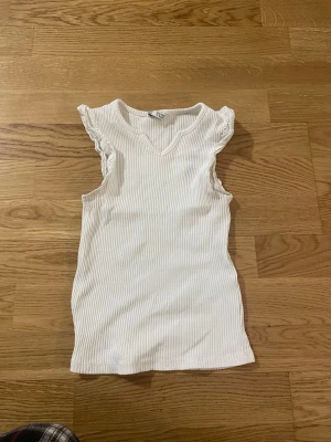 Vit ribbad ärmlös topp med volang - Säljer en vit ribbad topp från C med v-ringning och volangdetaljer vid axlarna. Toppen är ärmlös och har en slim passform som sitter snyggt på kroppen. Perfekt att styla med jeans eller kjol för en fräsch look.