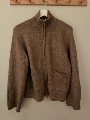 Beige ziptröja från Gina Tricot - Mysig beige ziptröja från Gina Tricot med hög krage och två stora fickor framtill. Tröjan är stickad i ett mjukt material och har ribbade detaljer vid krage, ärmslut och nederkant. Perfekt för lager på lager och en avslappnad look.