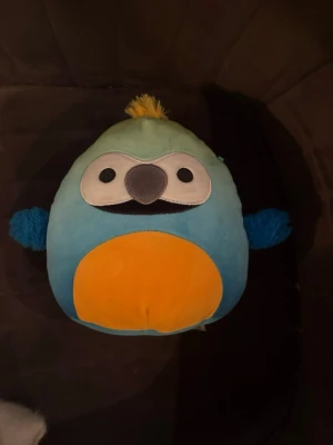 Blå Squishmallows papegoja - Mjuk och rund Squishmallows-gosedjur formad som en papegoja i blått, gult och vitt. Den har små blå vingar, gul mage och en söt näbb samt en gul tofs på huvudet. Perfekt som prydnad eller kramkompis.