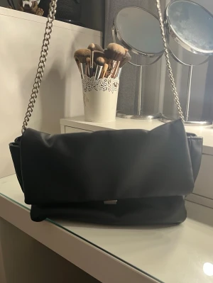 Svart axelväska med kedjerem - Stilren svart axelväska med bred klaff och grov silverfärgad kedjerem. Väskan har en mjuk, puffig form och är tillverkad i ett slätt material som liknar skinn. Perfekt för dig som gillar en enkel men trendig look.
