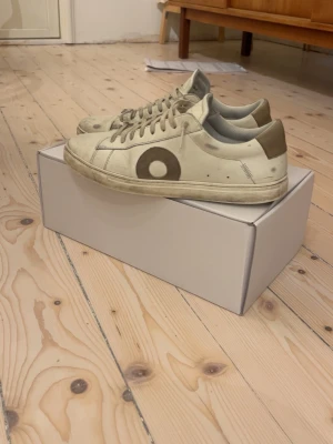 Oliver Cabell vita sneakers med bruna detaljer - Säljer ett par vita sneakers från Oliver Cabell med bruna detaljer runt vid framsidan av skon. Skorna har snörning, rund tå och är tillverkade i skinn. Klassisk låg modell som passar till det mesta.