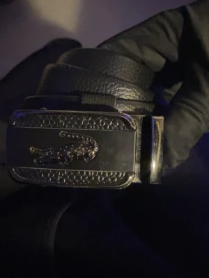 Svart Lacoste bälte med metallspänne - Snyggt svart bälte från Lacoste med ett stilrent metallspänne som har den klassiska krokodilloggan. Bältet är gjort i präglat läder och har en modern, elegant look som passar till många outfits. Perfekt accessoar för dig som gillar stilrena detaljer. Pris kan diskuteras vid snabb äffar. Först till kvarn!