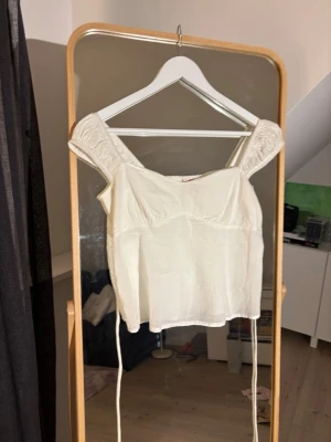 Vit linnetopp med knyt och puffärm - Supersöt vit linnetopp med puffiga axelband och knyt i ryggen. Toppen har smock baktill för skön passform och är lätt croppad. Perfekt till jeans eller kjol för en fräsch look.