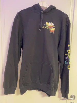 Svart hoodie med Svampbob-tryck - Cool svart hoodie från H&M med färgglatt Svampbob-tryck på bröstet och stort motiv med texten 'Stay Pretty' på ryggen. Har huva med dragsko, känguruficka och tryck på ena ärmen. Perfekt för dig som gillar streetwear och tecknade motiv.