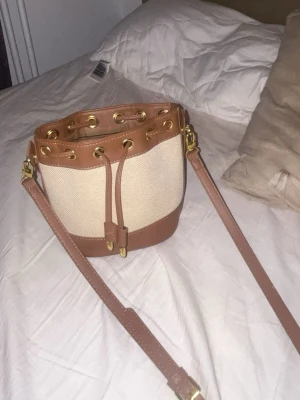 Beige och brun axelväska med snörning - Snygg beige och brun axelväska i bucket-modell med justerbar rem och guldfärgade detaljer. Väskan har snörning upptill och insida med randigt foder. Perfekt för dig som vill ha en stilren och trendig accessoar.