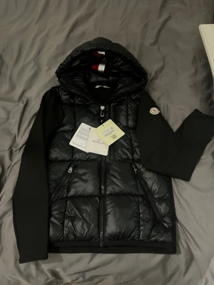 Svart pufferjacka från Moncler - Säljer en svart pufferjacka från Moncler med huva och två dragkedjeförsedda fickor. Jackan har Moncler-logga på ärmen och coola detaljer som snörning i huvan. Materialet är blankt syntet med quiltad look och släta ärmar. Perfekt för dig som vill ha en stilren och exklusiv jacka.